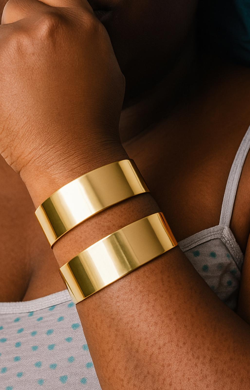 Close-up of a person wearing two shiny gold Space cuff bangle bracelets on their wrist, styled with a casual light blue polka dot top.