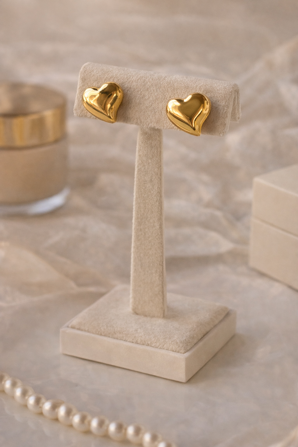 Sweetheart Gold Stud Earrings in polished gold, heart-shaped design displayed on a soft beige stand, styled with pearl necklace and luxury accessories background.