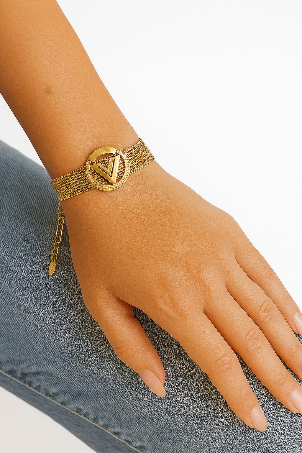 Close-up of a hand wearing a V-Mesh Gold Emblem Bracelet featuring a gold mesh band and a circular gold emblem with a V design, styled with casual denim.
