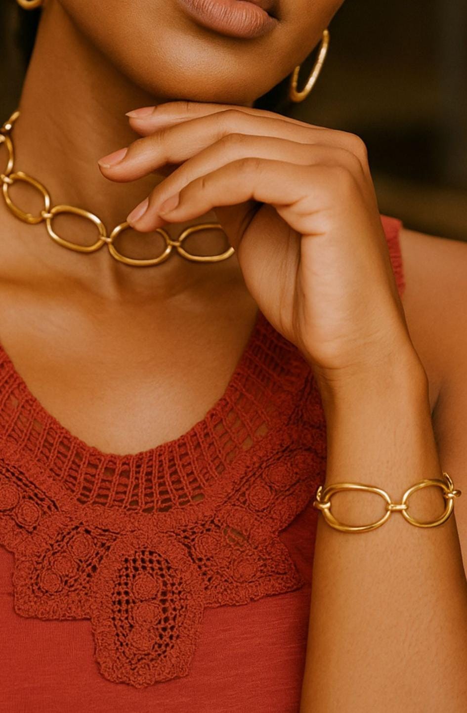 Close-up of a woman wearing a gold chain choker set featuring large oval links, paired with matching hoop earrings and bracelet, styled with a rust-colored knit top.