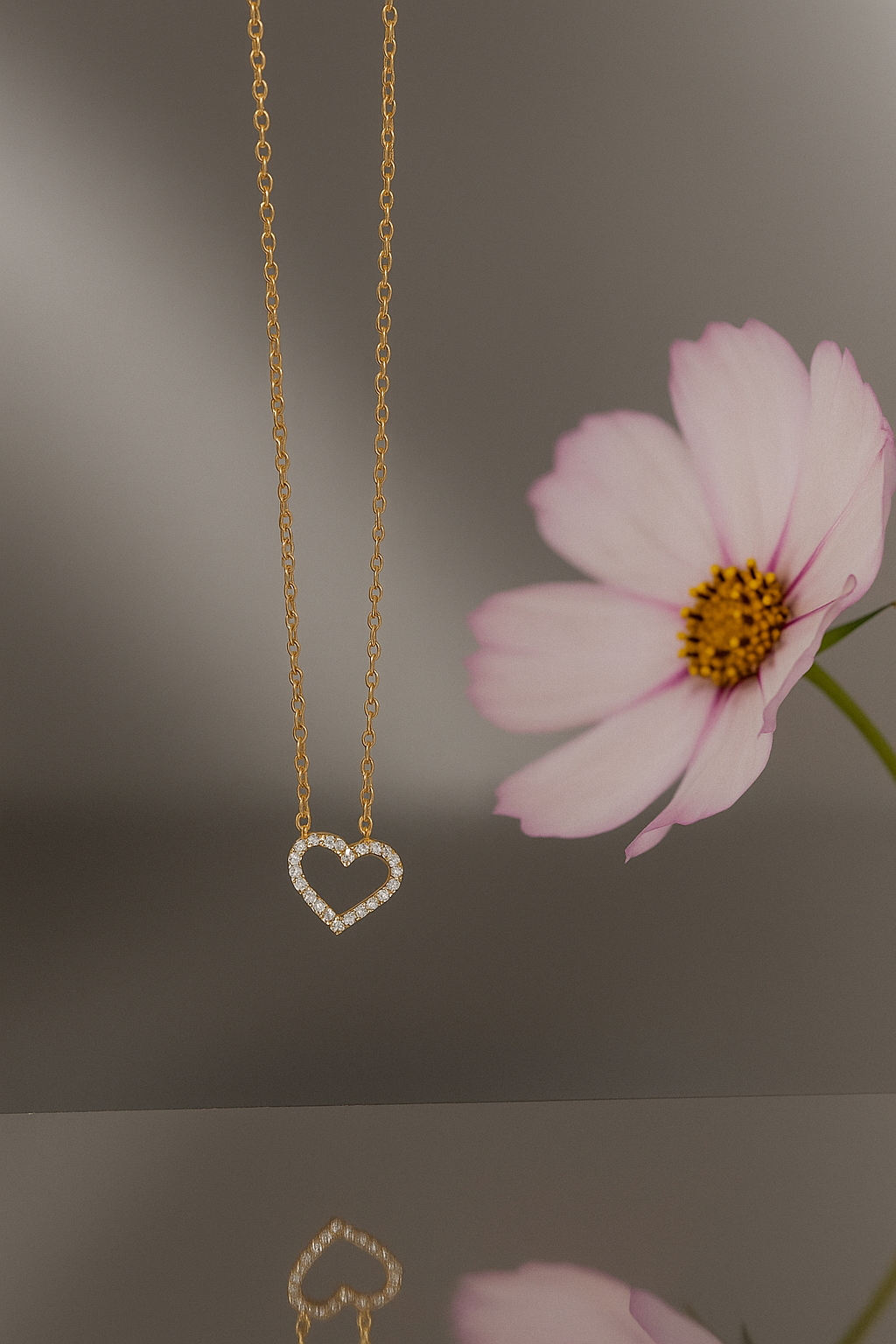 Delicate gold chain featuring a sparkling heart-shaped pendant adorned with small crystals, elegantly displayed next to a soft pink flower, showcasing the Stoned Heart Pendant Necklace.