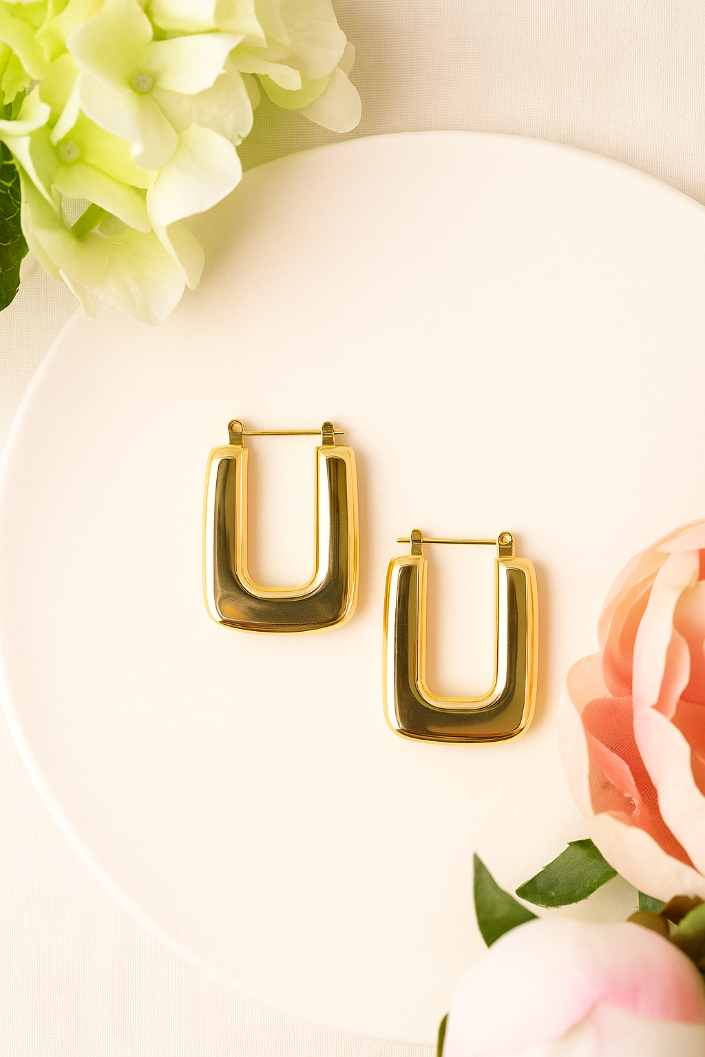 Pair of polished gold Square earring styled hoops on a white plate, elegantly surrounded by soft pink and white flowers for a luxury accessories presentation.