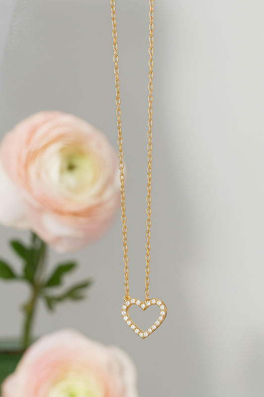 Stoned Heart Pendant Necklace featuring a delicate gold chain with a heart-shaped pendant adorned with small sparkling stones, styled against a soft floral background.