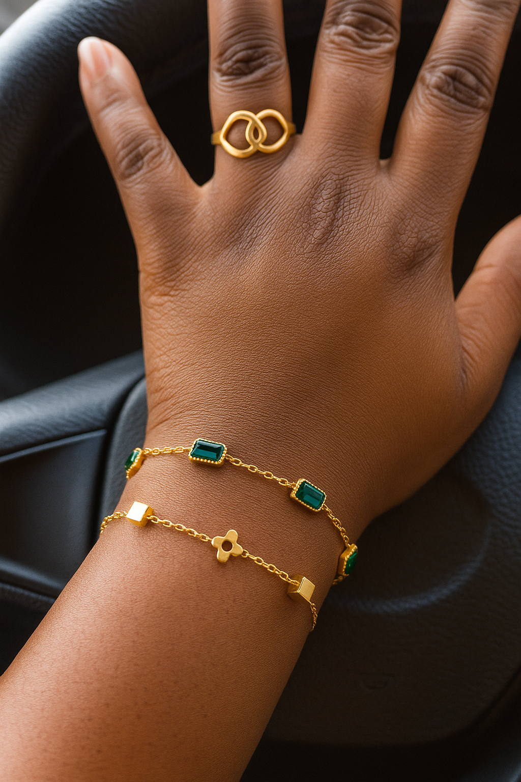 Close-up of a hand wearing a gold ring and two gold chain bracelets with green stones and geometric shapes, showcasing the Simple braclet stack on a dark background.