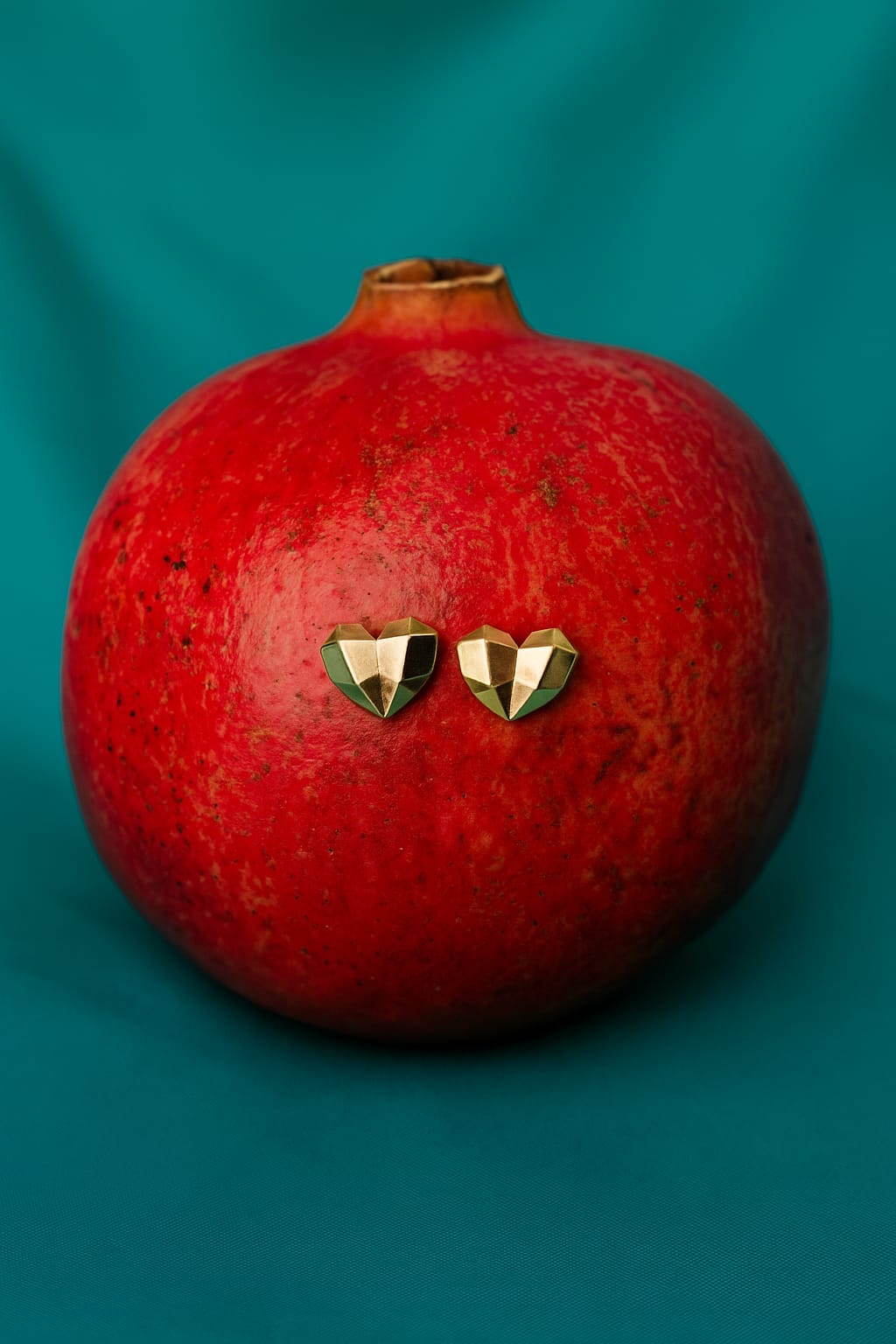 heart shaped stud earrings in gold placed on a red pomegranate against a teal background, showcasing luxury accessories with a geometric design