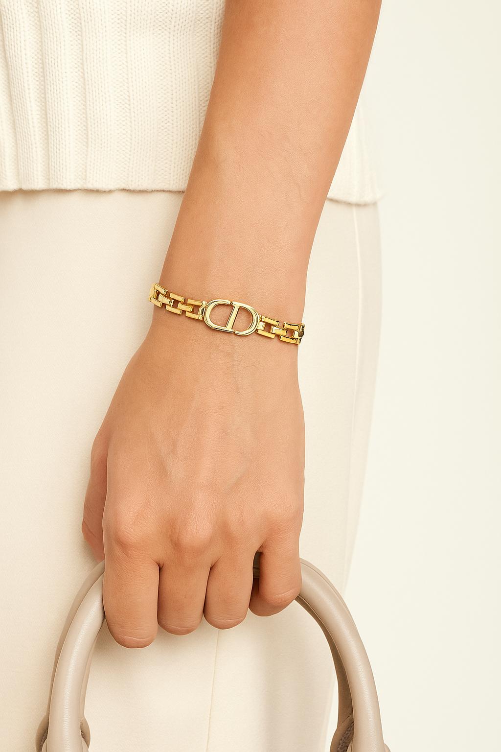 Close-up of a woman's wrist wearing a gold-tone CD inspired Braclet with interlocking chain links, styled with neutral-toned clothing and holding a beige handbag.