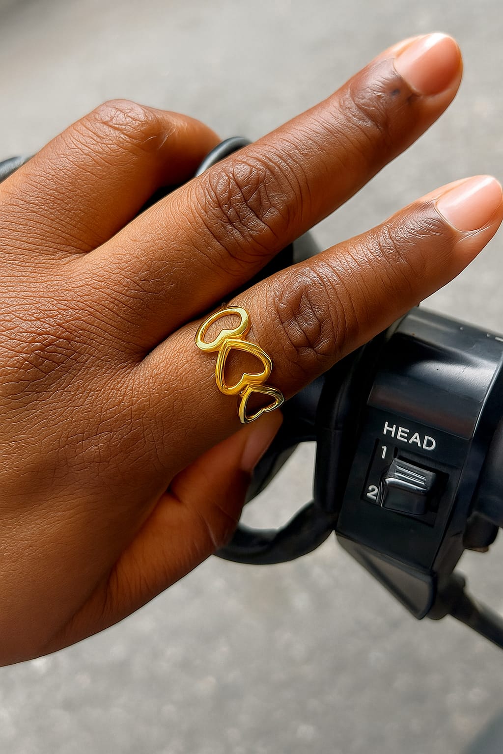 Close-up of a hand wearing a gold Multiple heart ring on the middle finger, resting on a black motorcycle handlebar with a control switch visible.