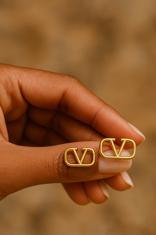 Close-up of a hand holding a pair of elegant gold V stud earrings against a soft brown background, showcasing luxury accessories with a minimalist design.