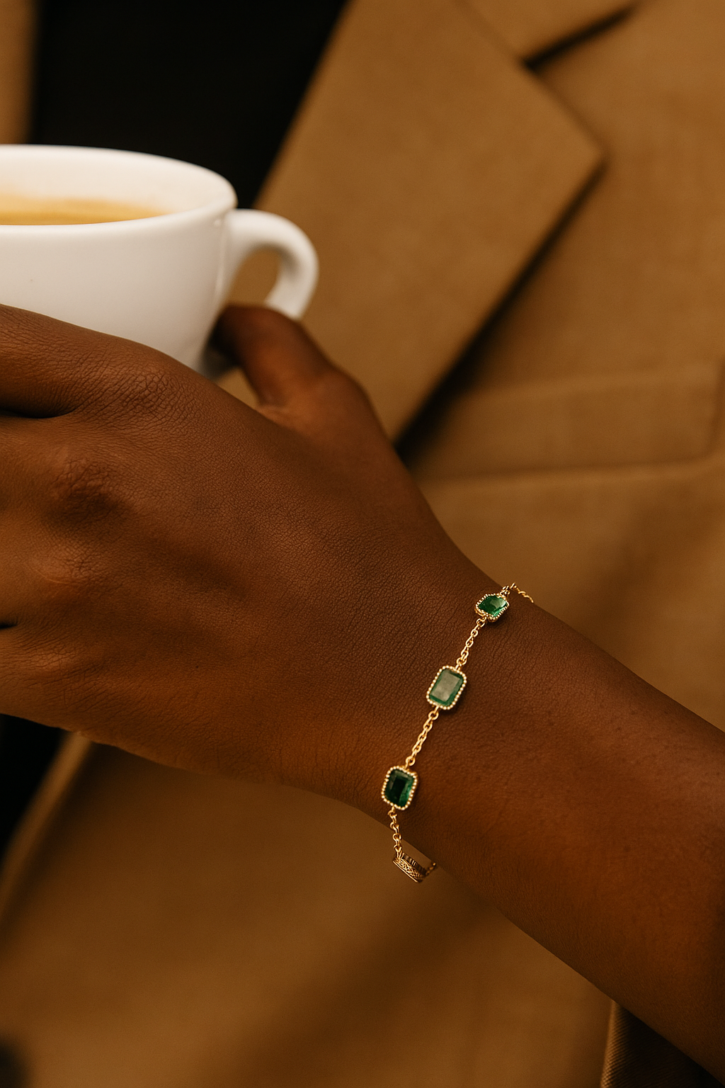 Close-up of a wrist wearing an Emerald braclet with green stones set in gold chain links, holding a white cup against a brown blazer background.