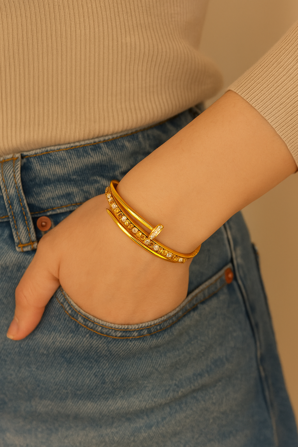 Close-up of a wrist wearing a gold diamond cut finish braclet paired with a light beige ribbed long sleeve top and blue denim jeans, showcasing luxury accessories.