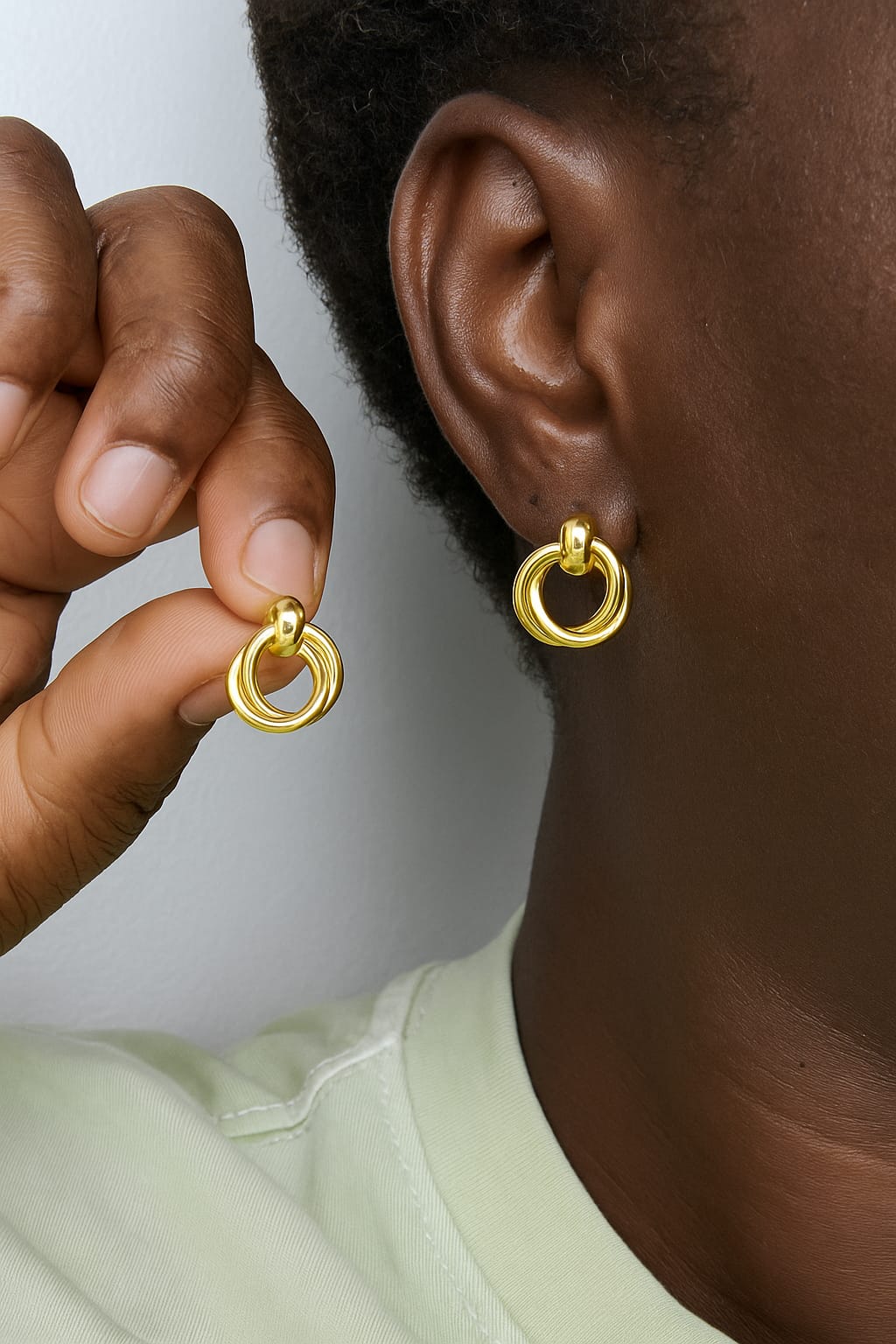 Close-up of a person wearing and holding a pair of shiny gold-tone Cute stud earrings, showcasing smooth circular hoops with a polished finish, styled with a light green top.