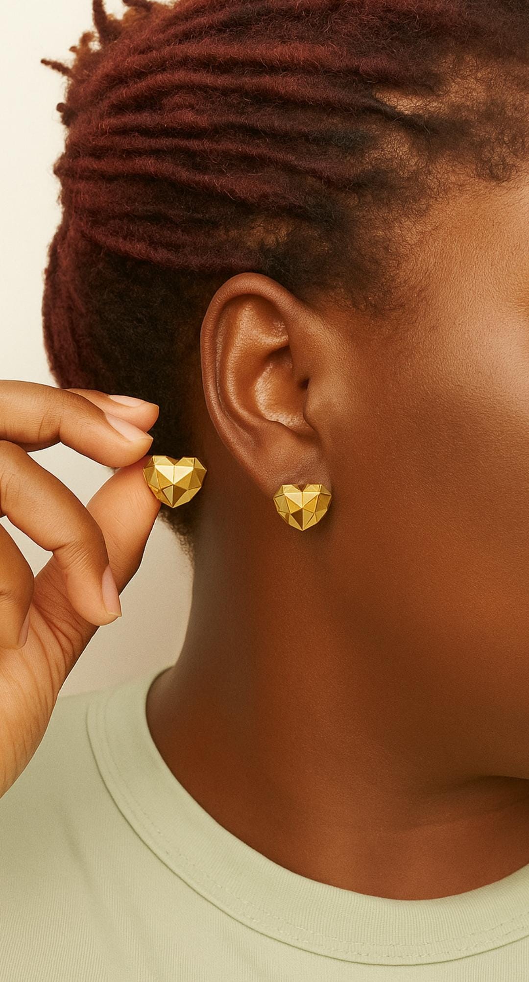 Close-up of a person wearing a pair of geometric gold heart shaped stud earrings, with one earring held between fingers, showcasing luxury accessories.