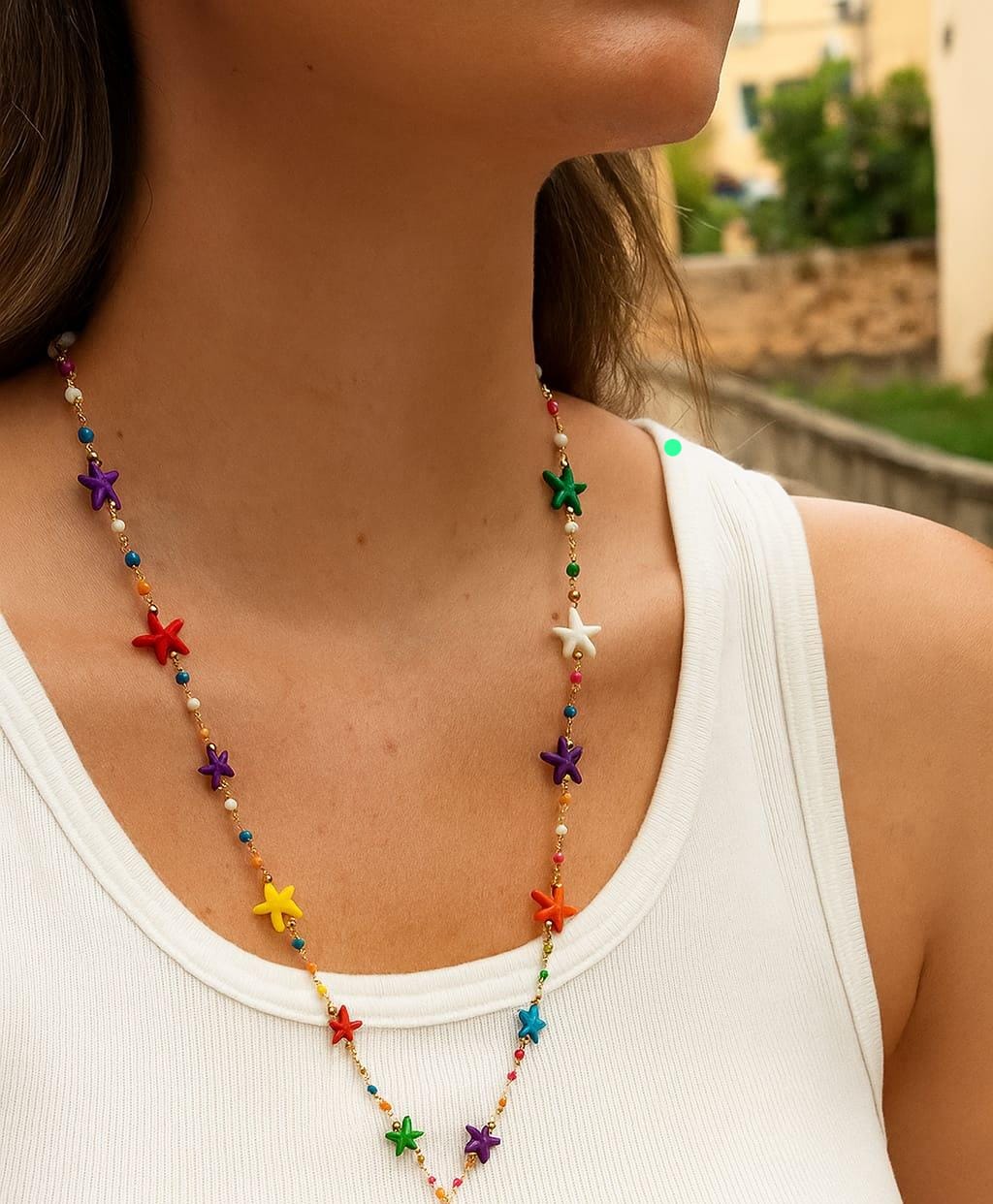 Close-up of a person wearing a multicolored starfish necklace featuring small gold accents and vibrant star-shaped beads against a white top outdoors.