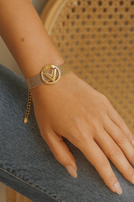 Close-up of a wrist wearing a V-Mesh Gold Emblem Bracelet featuring a textured gold mesh band and a circular gold emblem with a triangular design, styled with casual denim.