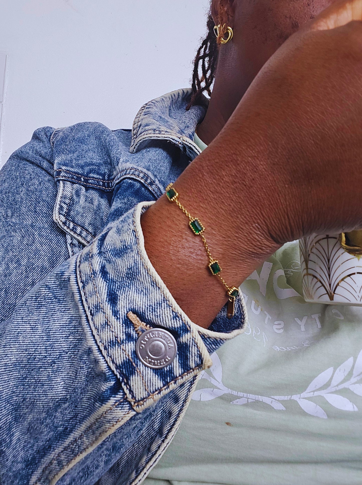 Close-up of a wrist wearing an Emerald braclet featuring small rectangular green stones set in a delicate gold chain, paired with a denim jacket and gold hoop earrings.
